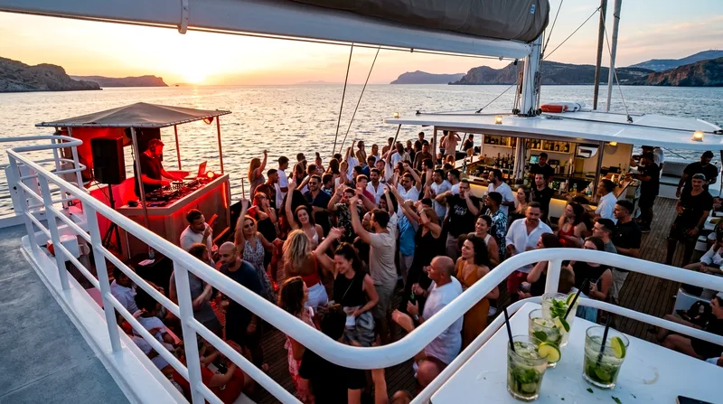 Large group of passengers dancing on a crowded catamaran deck near a DJ booth at sunset.